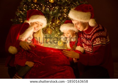Christmas Family in Santa Hat Looking on Gifts in Lighting Santa Bag. Happy Together open Presents under Xmas Tree on Merry Christmas Eve. Happy New Year Party Celebration