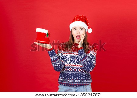 a young surprised woman with a gift Shoe in her hand in a sweater with snowflakes and a Santa Claus hat on a red background.