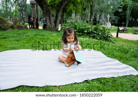 Funny black haired female toddler in light clothing sitting on blanket with soft toy and exploring picture book at summer park 
