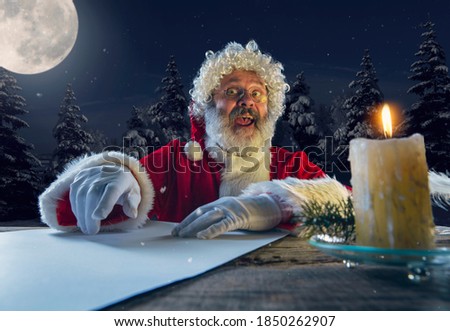 Emotional Santa Claus congratulating with New Year 2021 and Christmas. Man in traditional costume writing a letter, wish list lighted with candle and night forest on background. Winter, holidays.