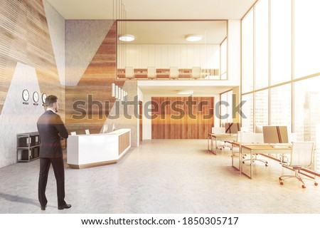 Young businessman standing in moder office with white and wooden walls, concrete floor, row of computer tables and reception desk. Toned image