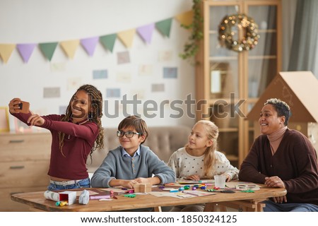 Waist up portrait of teenage African-American girl taking selfie with kids and teacher during art and craft class, copy space