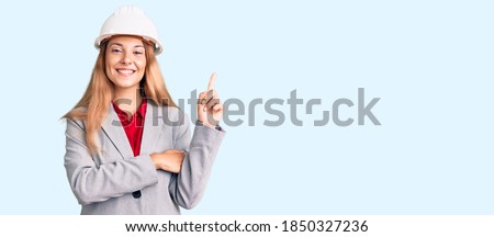 Beautiful young woman wearing architect hardhat with a big smile on face, pointing with hand and finger to the side looking at the camera. 