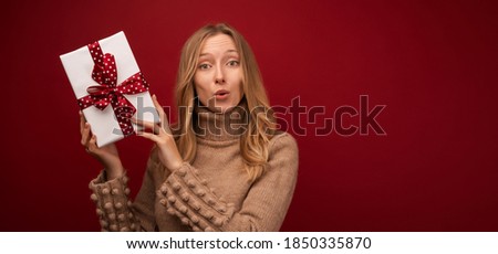 Image of charming young blonde woman in warm sweater smiling and holding gift with red ribbon. Studio shot red background. New Year Women's Day birthday holiday concept. Mock up copy space