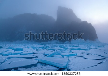 Scenic field of graphical cracked figured ice floes and mountain rock in fog on background, mystic winter morning landscape