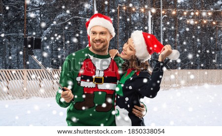 people, technology and holidays concept - happy couple in santa hats and sweaters taking picture with smartphone on selfie stick over skating rink and snow on background