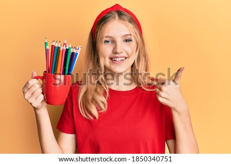 Beautiful young caucasian girl holding art colored pencils smiling happy and positive, thumb up doing excellent and approval sign 