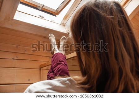 A young girl sits on Chrisla putting her foot on her leg against the background of an attic window in the attic of a wooden background