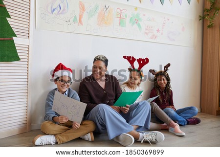 Full length portrait of female teacher sitting on floor with multi-ethnic group of kids holding pictures while enjoying art class on Christmas, copy space