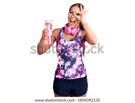 Young beautiful hispanic woman wearing sportswear holding water bottle smiling happy doing ok sign with hand on eye looking through fingers 