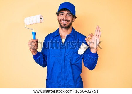 Handsome young man with curly hair and bear wearing builder jumpsuit uniform holding paint roller doing ok sign with fingers, smiling friendly gesturing excellent symbol 