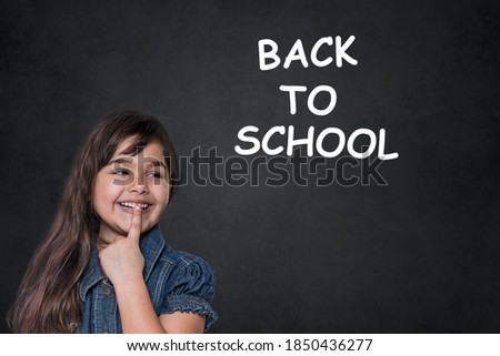 Cute tanned long haired  little girl is looking at the letters Back to School on the black chalkboard