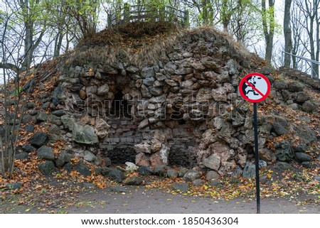 No climbing sign near the cliffs. Old ruins - potential shelter under the small hill. Dangerous area with rocks falling hazard. Autumn vibes and colors in the park at Tartu. Big red warning sign