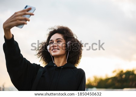 Cheerful athletic sportswoman in headphones taking selfie on mobile phone at city bridge