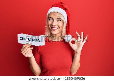 Young blonde girl wearing xmas hat holding paper with christmas word doing ok sign with fingers, smiling friendly gesturing excellent symbol 