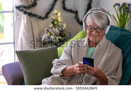Happy senior woman sitting at home on the sofa while listening to music with headphones while holding her smart phone. Christmas decoration on the background