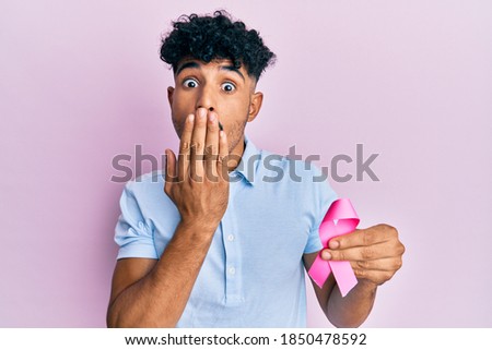 Young arab handsome man holding pink cancer ribbon covering mouth with hand, shocked and afraid for mistake. surprised expression 