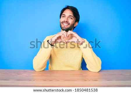 Handsome hispanic man wearing casual sweater sitting on the table smiling in love showing heart symbol and shape with hands. romantic concept. 
