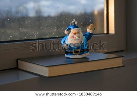 Christmas toy Santa Claus with a raised hand stands on a book against the background of a fogged window with rain drops.