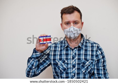 Man in checkered shirt show Costa Rica flag card in hand, wear protect mask isolated on white background. American countries Coronavirus concept.