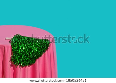 Artistic still life of a green cheerleader pompom on a circular table with a pink tablecloth and a very colorful blue background