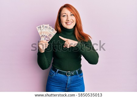 Beautiful redhead woman holding 500 mexican pesos banknotes smiling happy pointing with hand and finger 