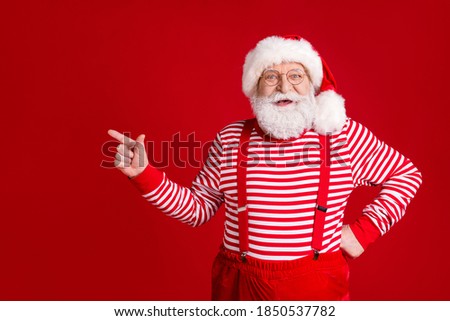 Photo of pensioner old man grey beard direct finger empty space offer best evergreen tree wear santa x-mas costume suspenders spectacles striped shirt headwear isolated red color background