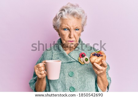 Senior grey-haired woman drinking a cup of coffee and eating bun skeptic and nervous, frowning upset because of problem. negative person.  Royalty-Free Stock Photo #1850542690