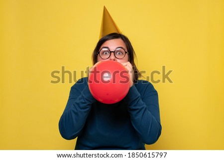 Caucasian woman in party hat blowing up red balloon preparing for birthday party.