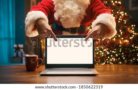 Santa Claus standing behind a desk and pointing at the laptop screen, Christmas and communication concept Royalty-Free Stock Photo #1850622319