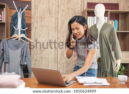 Young attractive female fashion designer leaning and use mobile talk to her customer on office desk, working with a laptop at home. Asian woman fashion designer working on her designs in the studio.