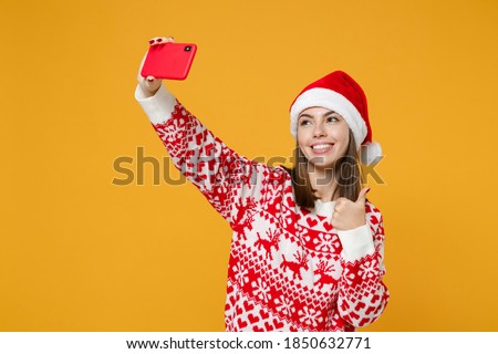 Smiling young Santa woman in sweater Christmas hat doing selfie shot on mobile phone showing thumb up isolated on yellow background, studio portrait. Happy New Year celebration merry holiday concept