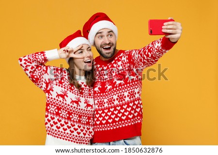 Excited young Santa couple friends man woman in sweater, Christmas hat doing selfie shot on mobile phone showing victory sign isolated on yellow background. Happy New Year celebration holiday concept