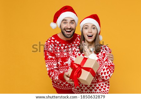 Surprised young Santa couple friends man woman 20s in red sweater Christmas hat hold present box with gift ribbon bow isolated on yellow background. Happy New Year celebration merry holiday concept
