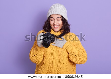 Woman photographer takes images with camera, has funny result, laughing happily and keeping palm on chest, wearing sweater, cap and mittens, posing against lilac background.