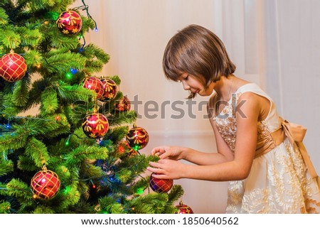 A little girl in a beautiful dress enthusiastically dresses up a Christmas tree