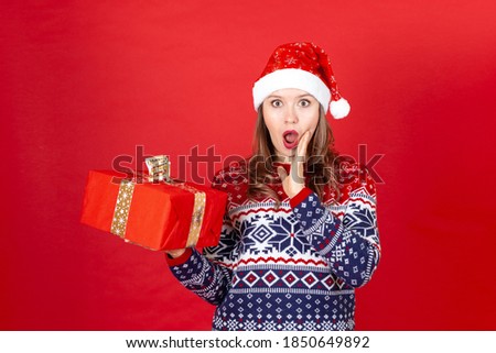 a surprised woman with a wide open mouth with a gift box in her hand in a sweater with snowflakes and a Santa Claus hat on a saturation background.