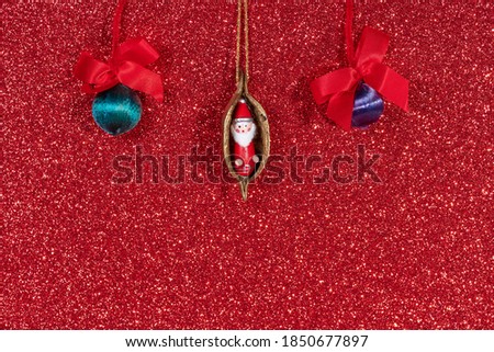 Image of a father Christmas tree decoration in a nut and two decorative balls with red ribbons against a red glitter background