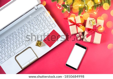 Online purchase of Christmas gifts using a laptop, phone and credit card on a red background with gifts and fir branches. New year, Christmas