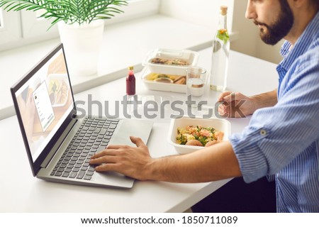Close-up side view of young man having takeaway meal during lunch break in office, using laptop computer, signing up for food delivery website and ordering some more food at discount online Royalty-Free Stock Photo #1850711089