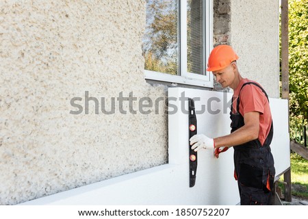 Insulation of the house with polyfoam. The worker is checking with the construction level the accuracy of the installation of polystyrene board on the facade. Royalty-Free Stock Photo #1850752207