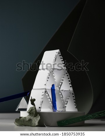 Modern still life, conceptual art: card pyramid from white paper, glass sticks, leaf on curved background, minimal colours. Instability, fragility