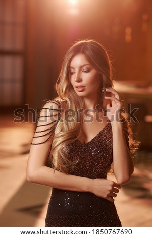 Elegant woman in long evining black dress with sequins standing and posing in luxury bar or restaurant interior with golden lightning