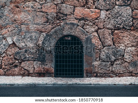 An arc door with a lattice in a stony wall of roughly hewn coquina stones and long dark sewer tunnel behind the entrance, an asphalt road and a small sidewalk with paving-stone in the foreground