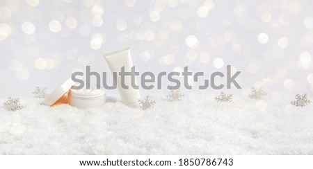 Winter skin care cosmetic products in snow and snowflakes on white background with bokeh lights. Opened face cream jar and hand creme or body lotion tube. Festive banner with copy space for text Royalty-Free Stock Photo #1850786743