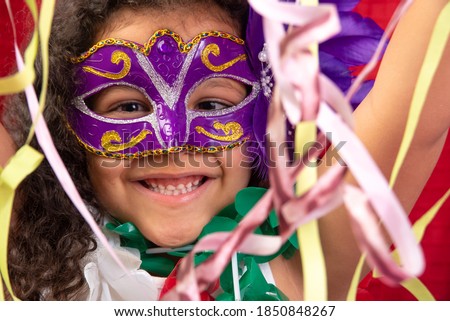 Small girl with mask and necklaces used in Brazil's carnival playing with streamers, red background, selective focus.