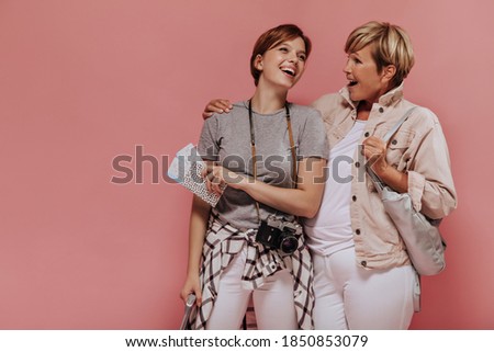 Stylish two ladies with cool short hairstyle in t-shirts and light trousers laughing and posing with tickets and camera on pink background..