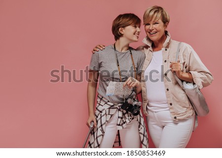 Cheerful two short haired women in white pants smiling, hugging and kissing with tickets, camera and bag on pink background..