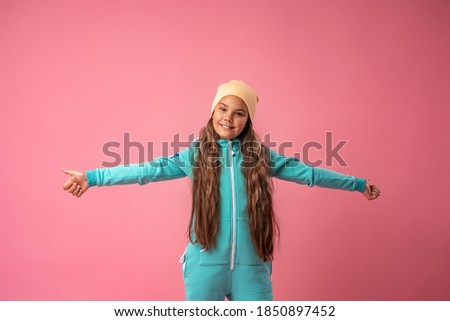 Teen girl in beanie, turquoise warm jumpsuit with hood and white sneakers stands on pink background with her arms outstretched to sides. Children's fashion comfortable children's clothing for autumn.