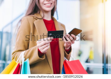 Cropped shot of elegant young woman using smartphone to check a balance on her credit card while standing with a bunch of colorful shopping bags in hands at the mall.